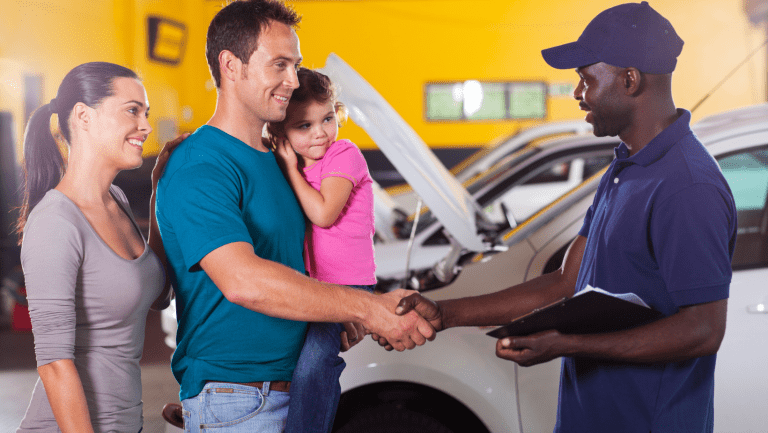 A family of 3 on the left shaking hands with a mechanic in a mechanic shop with a yellow background wall and cars in the background